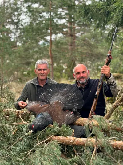 Two hunters smiling in the forest, holding a capercaillie with its tail fanned out