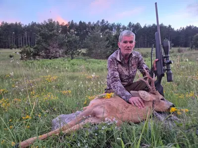 Un cacciatore accanto a un capriolo in un prato fiorito davanti a una pineta al crepuscolo