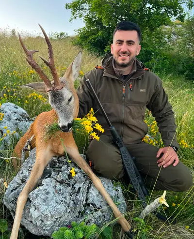 A young hunter in a green jacket kneeling beside a roe buck in a meadow of yellow wildflowers